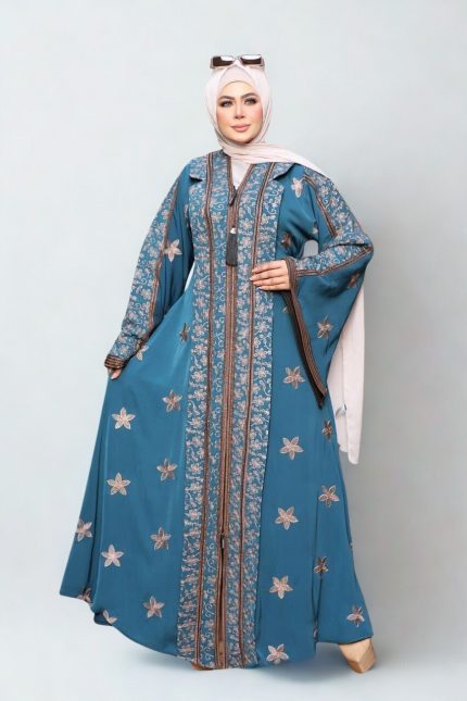 A woman wearing a teal embroidered abaya with floral patterns, styled with a beige hijab and sunglasses on her head, standing indoors in a chic setting